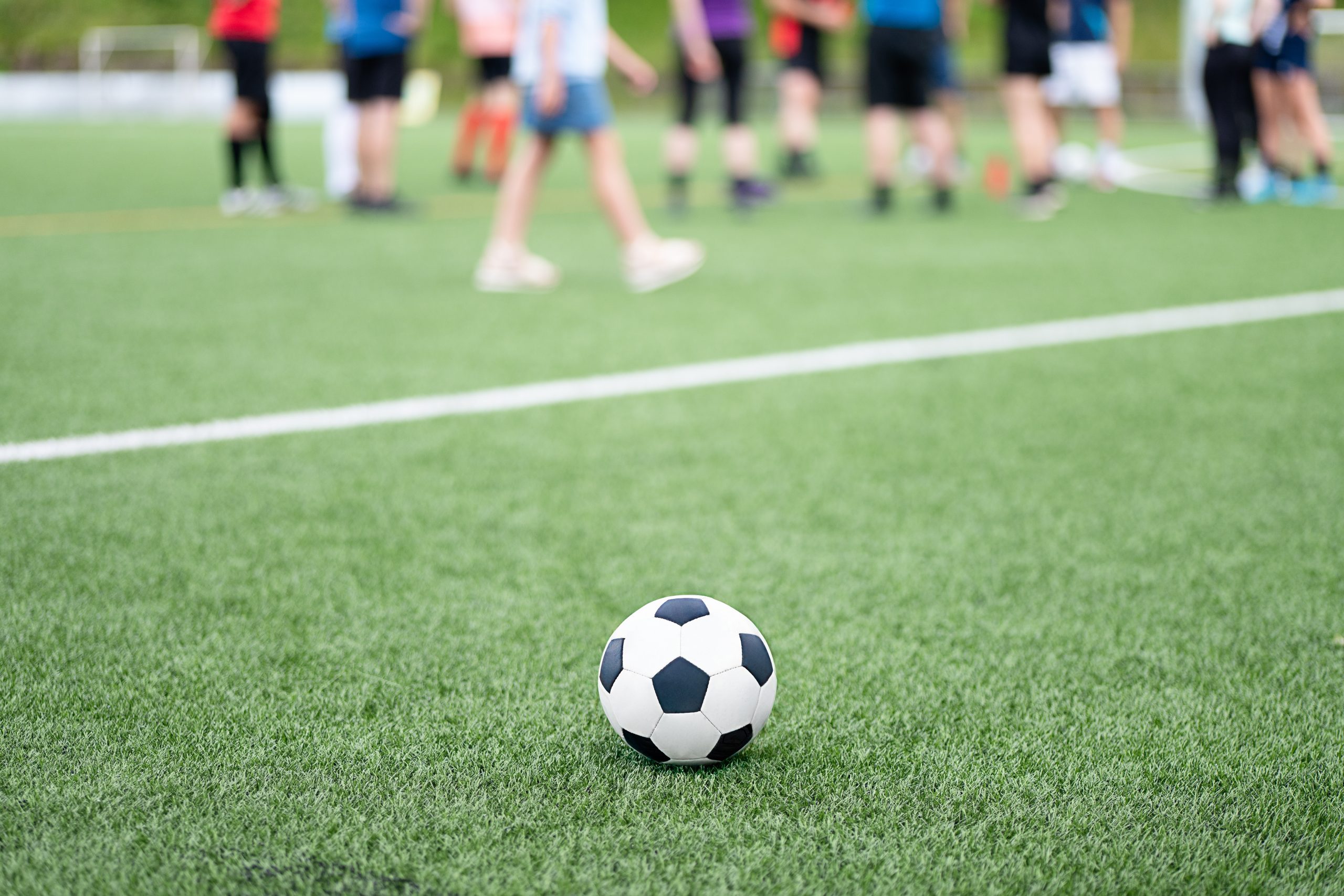 A soccer ball sits on the grass while players gather for practice, showcasing teamwork and energy during a vibrant day outdoors.